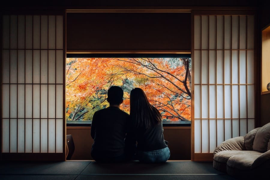 A loving young Asian couple sitting side by side by the window in a traditional Japanese style apartment, chatting and enjoying the beautiful nature Autumn scenics during the day